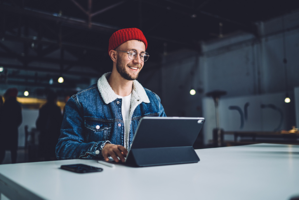 Young cheerful stylish bearded male student in warm clothes and glasses chatting on tablet in office sitting at white table