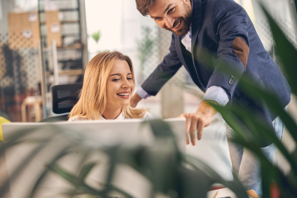 Beautiful female worker looking at computer and smiling while joyful man pointing at display